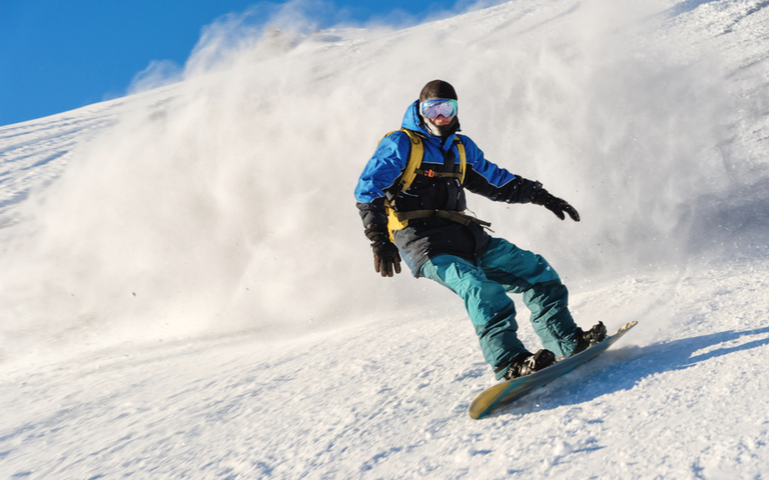 Snowboarder rolls on a snow-covered slope leaving behind a snow powder against the blue sky