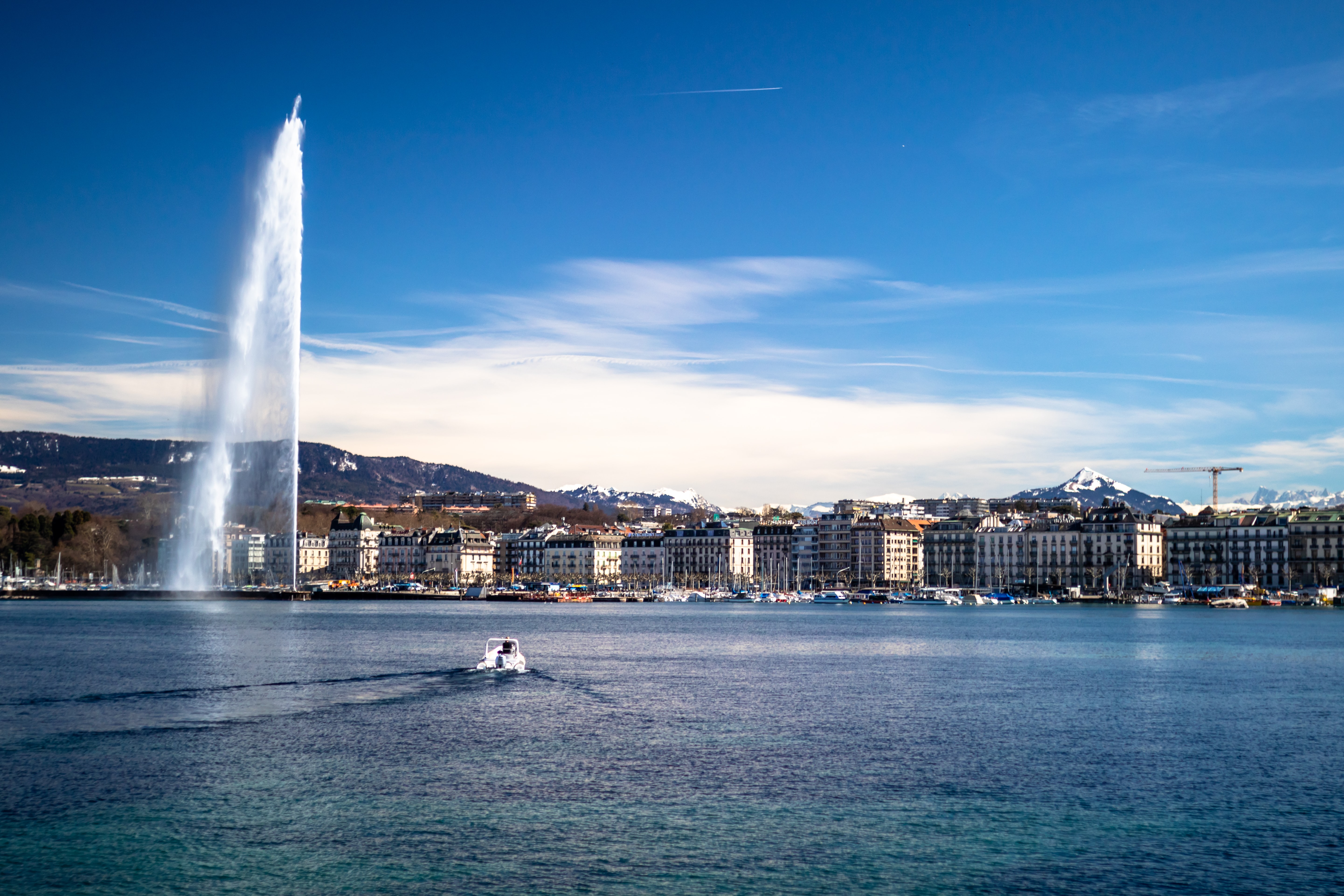 The beautiful city and the lake of Geneva with a boat, fountain, city and mountains in the distance


