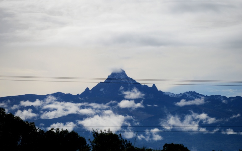 A scenic landscape of mount Kenya, a hiking mountain, with clouds and snow at its peak photographed in the morning from a distance with foggy grey skies and a forest in the view.
